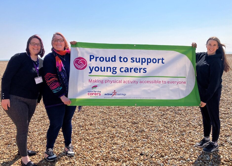 A photograph taken on the beach in Hastings. On the left are Rosie and Lisa from Care for the Carers' Young Carers Service with a member of Active Hastings on the right. They are holding a banner that reads 'Proud to be supporting young carers'