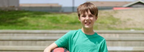 A young boy in a green tshirt, holding a football outside, facing the camera.